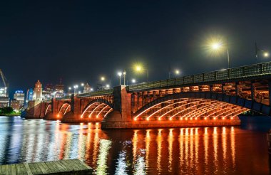 Boston 'daki Longfellow Köprüsü' nün gece görüşü turuncu ışıkla parıldıyor, Charles Nehri 'nin üzerinde dalgalanan yansımalarla ve arka planda şehrin ufkuyla birlikte.