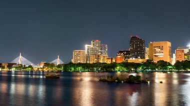 Zakim Köprüsü ve Boston şehir merkezinin panoramik gece manzarası Charles Nehri 'ne yansıyor. Parlayan şehir ışıkları ve kıyıda ağaçlar var. Massachusetts, ABD