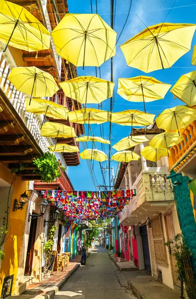 Kolombiya, Cartagena 'da renkli koloni binalarıyla dolu dar bir yaya sokağının üzerinde parlak sarı şemsiyeler ve uluslararası bayraklar asılı. Canlı dekorasyonlar açık mavi gökyüzünün altında, balkonları ve caddeleri olan neşeli bir atmosfer yaratıyor.