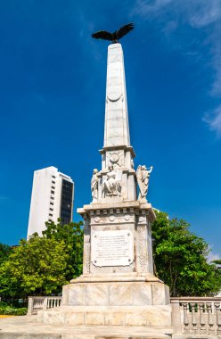 Kolombiya, Cartagena 'daki Centenario Parkı' nda kartal desenli mermer bir dikilitaşın üzerinde duruyor. Klasik heykeller tabanı süslüyor ve açık mavi gökyüzüne karşı anıtı çevreleyen yemyeşil yeşillikler var.