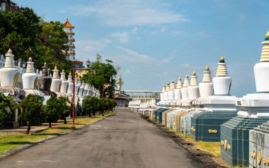 Beyaz stupaların simetrik sıraları, Tayvan 'ın Kaohsiung kentindeki Fo Guang Shan Manastırı' nda berrak mavi gökyüzünün altındaki ağaçların arasından görünen geleneksel bir Çin tapınağı ile sessiz bir patikadır.