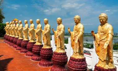 Tayvan 'ın Kaohsiung kentindeki Fo Guang Shan Manastırı' nın geleneksel kırmızı kiremitli koridoruna dizilmiş altın Buda heykelleri. Şekiller, dekore edilmiş bir çatının altında, canlı mavi bir gökyüzünün altında, huzurlu pozlarda duruyor.