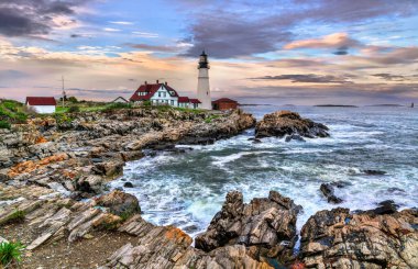 Portland Head Light dramatik okyanus dalgaları ve renkli akşam gökyüzü çerçeveli Maine, Cape Elizabeth 'in kayalık kıyılarında belirgin bir şekilde duruyor. Tarihi deniz feneri ve kırmızı çatılı binaları doğal granitle çok güzel tezat oluşturuyor.