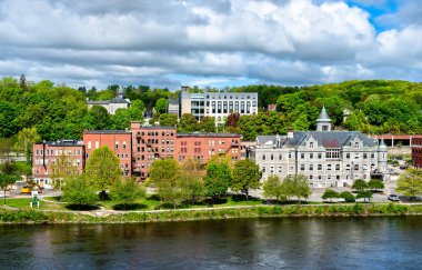 ABD 'nin Maine kentindeki tarihi Augusta şehir merkezine doğru Kennebec Nehri' nin karşısına bakın, Eski Postane, kırmızı tuğlalı binalar ve parçalı bulutlu gökyüzünün altında ağaçlarla kaplı tepeler yer alıyor.