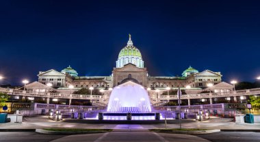 Harrisburg 'daki Pennsylvania State Capitol' ün gece manzarası, aydınlatılmış kubbe ve ön plaza çeşmesi sergileniyor. Güzel Sanatlar mimarisi, derin mavi gökyüzünün altında çarpıcı bir aydınlatma ile vurgulanır.