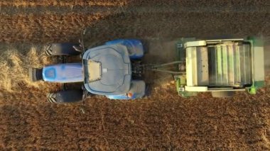 Üst manzarada, doli yukarı doğru hareket ediyor, traktörü takip ederek merdaneyi çekiyor, kamışı yuvarlayan ve tarımsal alanın üzerine yuvarlanmış yuvarlak bir balya püskürten makine..