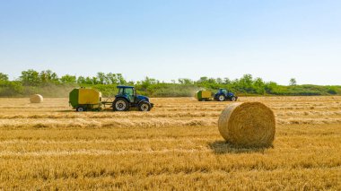 Hasattan sonra saman balyasının üstünde, arka plandaki traktör samanı çekiyor, kamışı yuvarlayan ve tarımsal alana tüküren makine..
