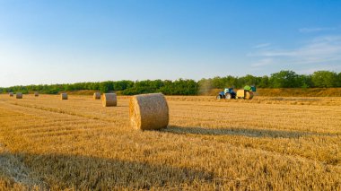 Hasattan sonra saman balyasının üstünde, arka plandaki traktör samanı çekiyor, kamışı yuvarlayan ve tarımsal alana tüküren makine..