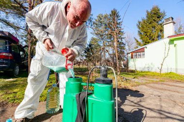 Farmer is preparing mixture of water and botanical solutions of pesticide in plastic knapsack sprayer to spray fruit trees in orchard to protect them with chemicals from fungal disease or vermin. clipart