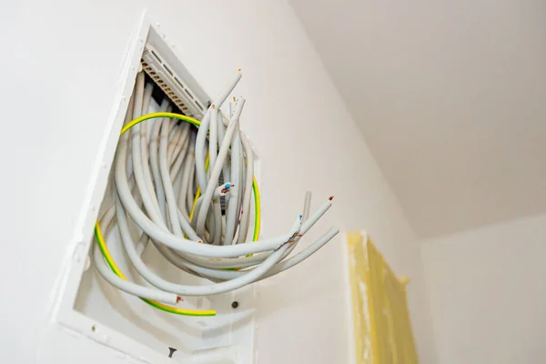 Bundle of various cables hang from an open plastic fuse switch box mounted on the plaster wall, electric power meter covered with duct tape installed for measuring power usage.
