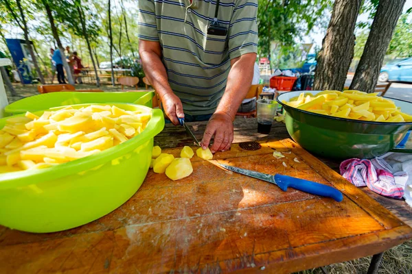 An elderly farmer cooker cuts potatoes using knife into slices on the wooden board, prepares it for cooking at the outdoor. Bowls are full of cut vegetable