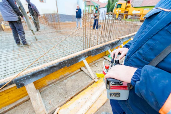 Operator, worker is holding industrial remote, wireless, console control with joystick to directing pump tube on right direction, pouring layer of concrete in building foundation, construction site.