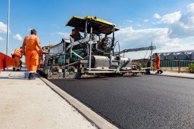 View on machine for laying asphalt, spreading layer of hot tarmac on prepared ground a few workers are working around.  