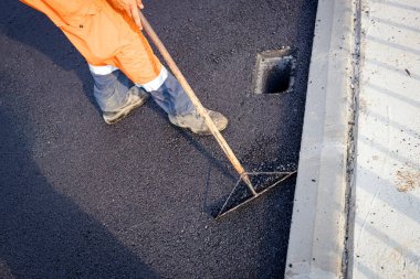 Above view on worker that uses rake to level, set up layer of fresh tarmac to right measures, flattens the hot asphalt around the quadratic rainwater hollow.