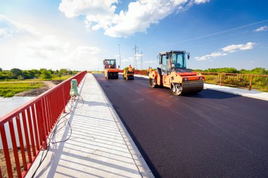 View on several steamrollers that are flatting fresh asphalt, spreading layer of hot tarmac on prepared ground at the bridge under construction.