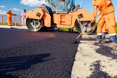 Workers are using rakes to level, set up layer of fresh tarmac to right measures, steamroller is flatting fresh asphalt .