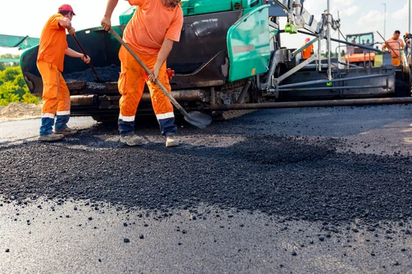 View on machine for laying asphalt, spreading layer of hot tarmac on prepared ground a few workers are using shovels to level it.