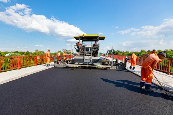 View on machine for laying asphalt, spreading layer of hot tarmac on prepared ground a few workers are using shovels to level it.