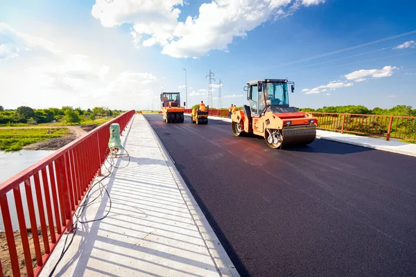 View on several steamrollers that are flatting fresh asphalt, spreading layer of hot tarmac on prepared ground at the bridge under construction.