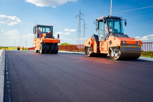 Several steamrollers that are flatting fresh asphalt, spreading layer of hot tarmac on prepared ground at the bridge under construction.