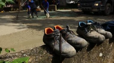 Old worn out safety shoes lined, placed on small wall near construction site as workers in boots are pouring concrete and using rake to spreading, leveling fresh cement.