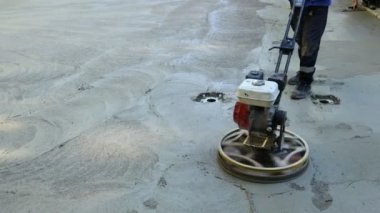 Worker using self-leveling power trowel machine with gas engine has wheels with pads, plate and he is driving it in circles smoothing surface on concrete slab.