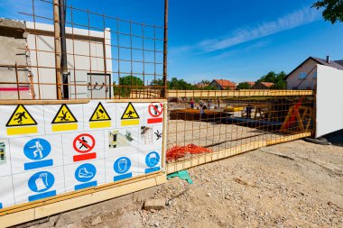 Board with safety regulations is standing in front fenced edifice under construction. Entrance, gate made of square reinforcement, on building site, restricted area, for the authorized personnel only.  clipart