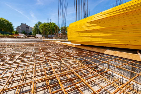 Perspective view on rusty, quadratic reinforcement, armature, mesh placed in foundation for concrete strength of residential building that is under construction. Pile of painted yellow wooden planks.