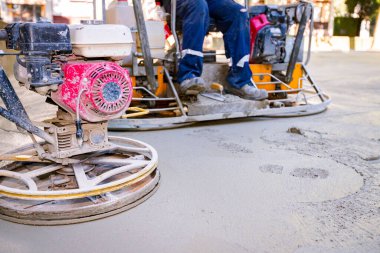 Two types of self-leveling power trowel machine with gas engine have wheels with pads, plate and he is driving it in circles smoothing surface on concrete slab.