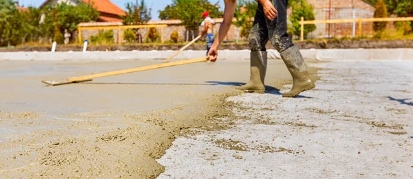 Rigger in rubber boots, worker is using wooden handmade handcraft tool to leveling fresh concrete after pouring in building foundation.