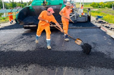 Zrenjanin, Voyvodina, Sırbistan - 8 Haziran 2021: Birkaç işçi asfalt seviyesini yükseltmek için kürek kullanıyor, arkalarında inşaat halindeki köprüdeki hazır yol zeminine sıcak asfalt tabakası yaymak için kullanılan bir makine bulunuyor.