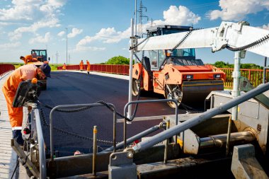 View on machine for laying asphalt, spreading layer of hot tarmac on prepared ground a few workers are working around.  