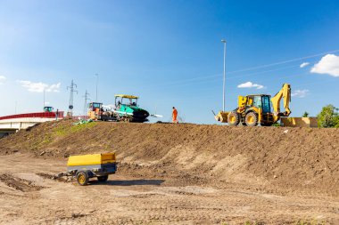 Spreader machine lays, pours hot asphalt, laying asphalt. Pneumatic compressor and excavator parked on construction zone. Road works on unfinished bridge. 