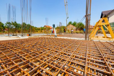 Paslı dikdörtgen çelik yığını inşaat alanına yerleştirilmeye hazır. Arka plandaki işçiler temel inşaata beton döküyorlar.