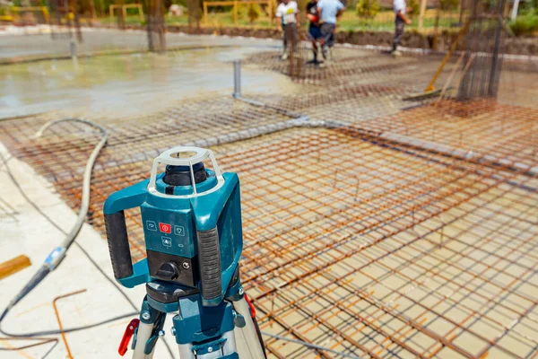 Total center device with laser for leveling other devices to level construction site. Workers measure and control level of concrete with other appliance as pouring into building foundation.