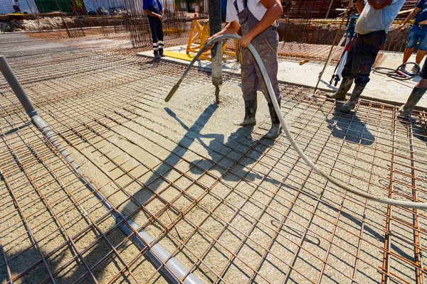 Worker is using vibration power tool, compactor, for compacting fresh liquid concrete, flowing slowly among square reinforcement in the base of new building