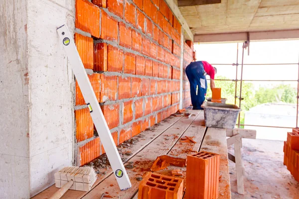 Spirit level for control measurements leaned to concrete pillar and a few red ceramic blocks placed on wood platform on construction site. Worker builds partition wall with blocks and mortar