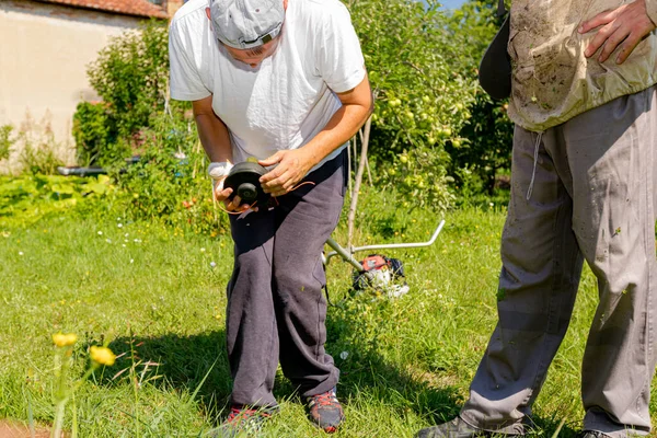 包帯の傷んだ手の修理トリマーを持つ修理工は トリマーヘッドコイルの文字列を調整し 果樹園や食前酒で草を刈るために全体的に保護で彼の友人養蜂家を助けます 庭道具の整備 — ストック写真