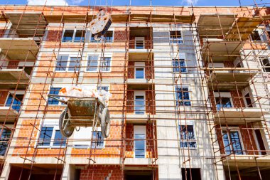 Vinçin altından çekilen ve endüstriyel atıklarla dolu bir el arabasına konan, buruşmuş selofan, arka planda yapı iskelesi olan bir yapı var..