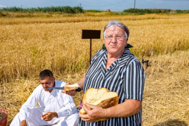 Muzlja, Voyvodina, Sırbistan, - 02 Temmuz 2022; Geleneksel olarak buğday hasadı. Yaşlı kadının portresi, büyükanne ekmek tutuyor ve kesiyor. Çiftçiler kahvaltı yapıyor, hasat için hazırlanıyorlar. 