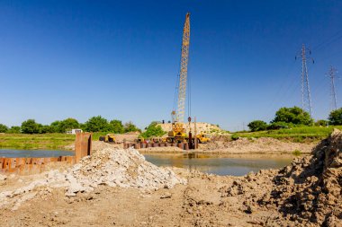 Toprak tepesi, nehir kıyısı boyunca inşaat alanında köprü temeli olarak inşa edilmiş metal yığınları. Suyun karşısında sarı renkli, pnömatik çekiçli taşınabilir vinç var..