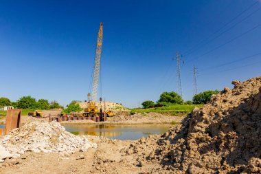 Toprak tepesi, nehir kıyısı boyunca inşaat alanında köprü temeli olarak inşa edilmiş metal yığınları. Suyun karşısında sarı renkli, pnömatik çekiçli taşınabilir vinç var..