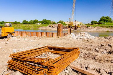 İnşaat alanına ikinci dereceden paslı destek çubukları yerleştirilmiş. Nehir kıyısındaki köprü temeli için monte edilmiş metal yığınları arka planda pnömatik çekiçli vinç bulunuyor..