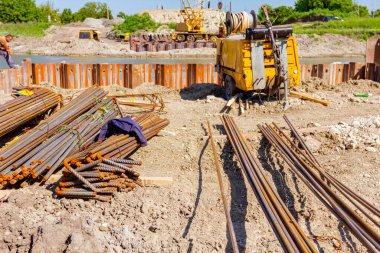 İnşaat alanına park edilmiş seçilmiş destek çubukları ve sarı kompresör paketleri. Arka planda nehir kıyısındaki köprü temeli için monte edilmiş metal yığınları pnömatik çekiçli vinçtir..