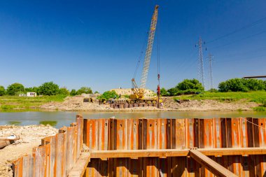 İnşaat alanındaki köprü temeli için nehir kıyısı boyunca inşa edilmiş paslı metal yığınları. Suyun karşısında sarı renkli, pnömatik çekiçli taşınabilir vinç var..