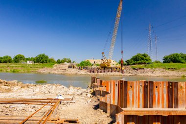 İnşaat alanındaki köprü temeli için nehir kıyısı boyunca inşa edilmiş paslı metal yığınları. Suyun karşısında sarı renkli, pnömatik çekiçli taşınabilir vinç var..