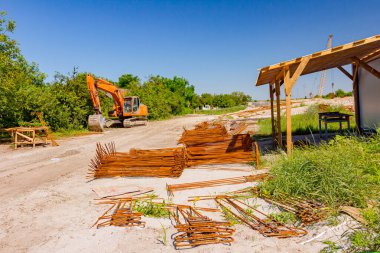 İnşaat alanında yerde paslı destek çubukları parçaları olan Alfresco doğaçlama depo, arka planda park edilmiş kazıcı var..