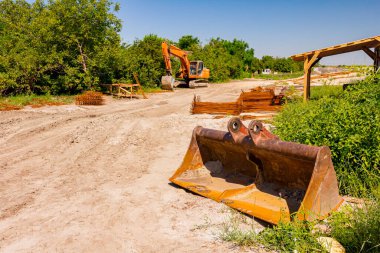 Kazıcının çalışma aleti eski paslı kova, el yapımı depo. İnşaat alanında parçalanmış paslı destek çubukları var. Park etmiş kazıcı arka planda..