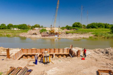 Köprü temelleri metal yığınları, kompresör, inşaat alanında armatür, jeodezist elektronik dengeleyici ile çalışıyor. Nehrin karşısında, hava basınçlı taşınabilir vinç var..