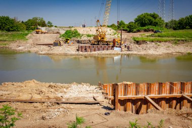 İnşaat alanında köprü temeli olarak inşa edilmiş nehir kıyısı boyunca metal yığınları var. Suyun karşısında sarı renkli, pnömatik çekiçli taşınabilir vinç var..